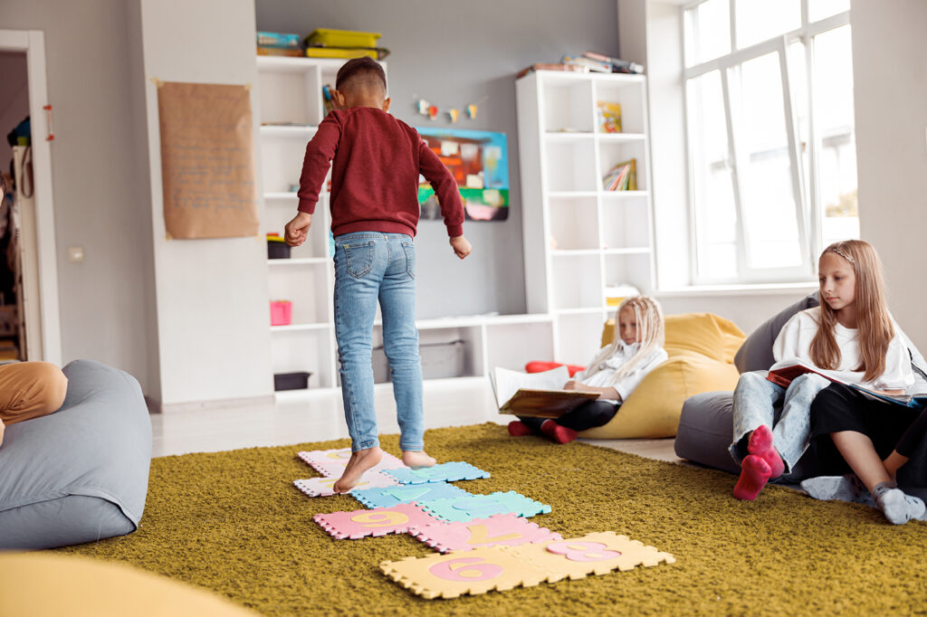 Boy jumping on a puzzle with numbers while girls resting on poufs in room in school. Education, school concept