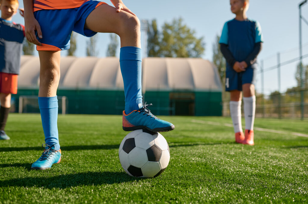 Young soccer player holding boots on ball cropped shot. Kids team practicing soccer on grass venue